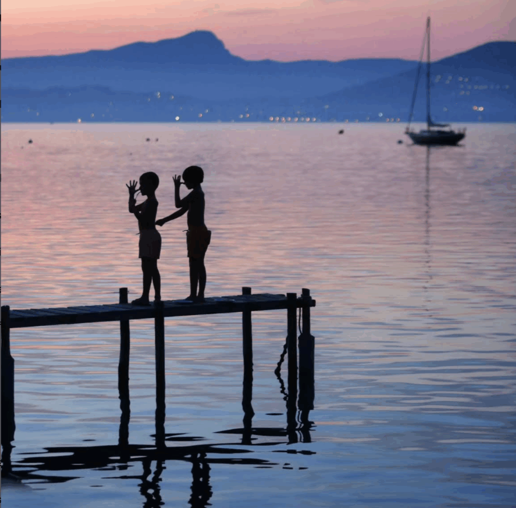 Deux enfants debout sur un ponton en bois au bord d’un lac au crépuscule, se faisant face dans une lumière rose-violette, avec un voilier et des montagnes en arrière-plan