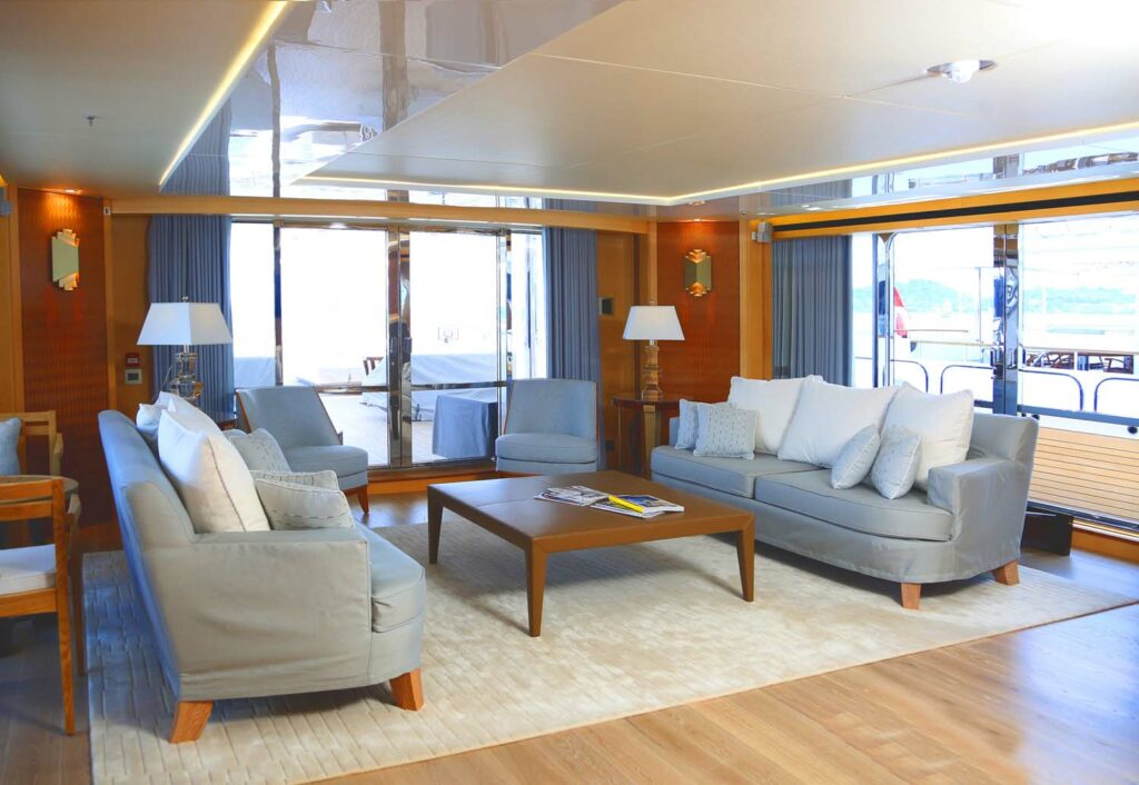 Salon lumineux et élégant à l’intérieur d’un yacht, décoré de fauteuils et canapés clairs autour d’une table basse en bois, avec de grandes baies vitrées donnant sur l’extérieur