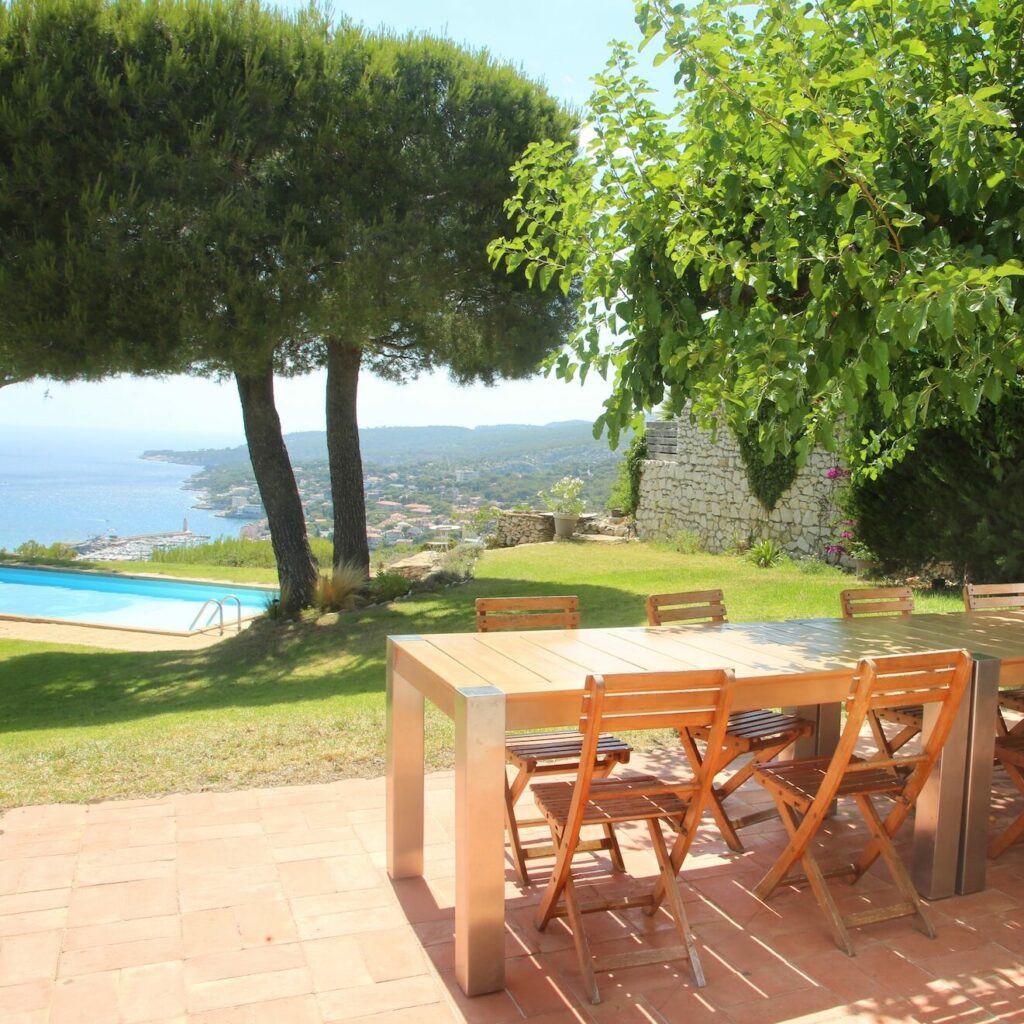 Terrasse extérieure ensoleillée avec une grande table en bois et des chaises, donnant sur une pelouse avec vue panoramique sur la mer et les collines, près d’une piscine