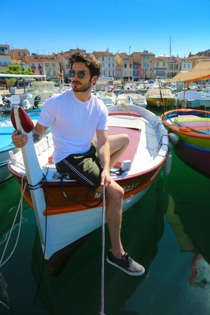 Jeune homme assis sur le bord d’un petit bateau amarré dans un port ensoleillé. Il porte un t-shirt blanc, un short foncé et des lunettes de soleil, avec une ville côtière en arrière-plan